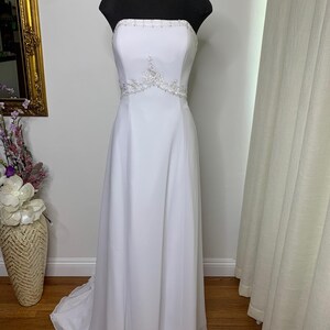 May include: A strapless white wedding dress on a black mannequin. The dress features delicate beaded embellishments around the bust and waist. The skirt flows to the floor with a slight train. The dress is displayed in a well-lit room.