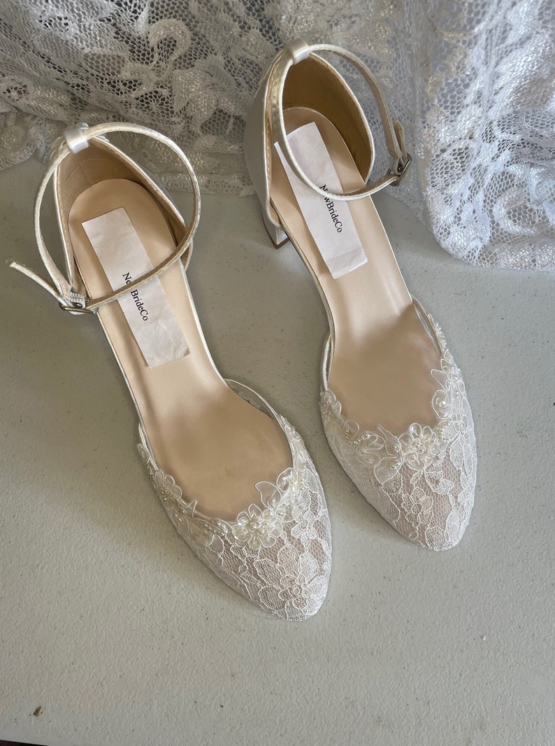 May include: A pair of ivory-colored bridal shoes with a low heel and ankle strap. The toe of each shoe is adorned with delicate lace and small pearl-like beads. The shoes have a label that reads "New BrideCo".