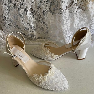 May include: A pair of white bridal shoes with a low heel. The shoes feature a delicate lace overlay on the toe and sides, adorned with small pearl accents. Ankle straps and a block heel complete the design. The label reads "New BrideCo".