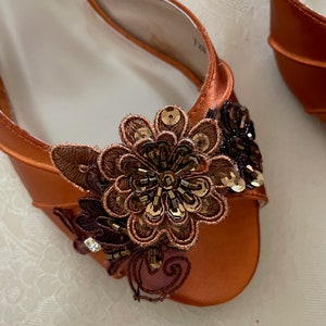 May include: An orange satin open-toe shoe featuring a decorative floral applique on the front. The applique consists of brown and copper-toned embroidery, dark brown beads, and metallic sequins arranged in a layered, multi-petaled flower design.