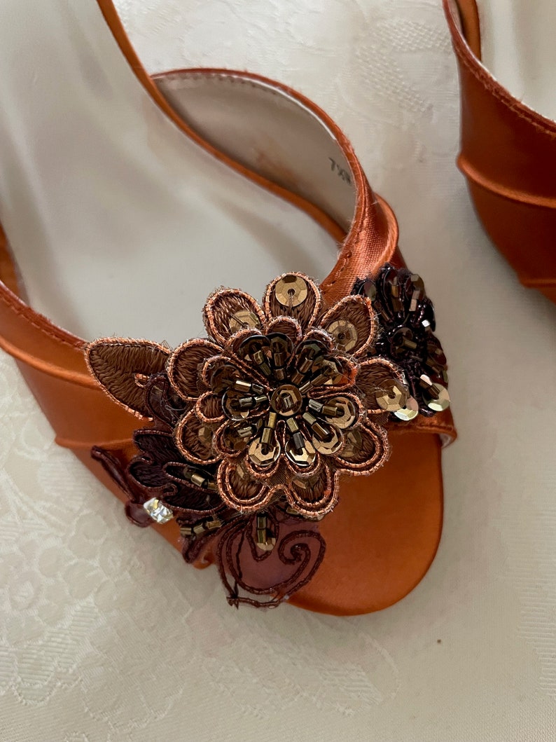 May include: An orange satin open-toe shoe featuring a decorative floral applique on the front. The applique consists of brown and copper-toned embroidery, dark brown beads, and metallic sequins arranged in a layered, multi-petaled flower design.