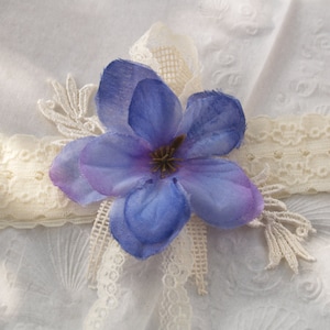 May include: A white lace garter with a blue flower accent. The flower has a purple center and is made of fabric.