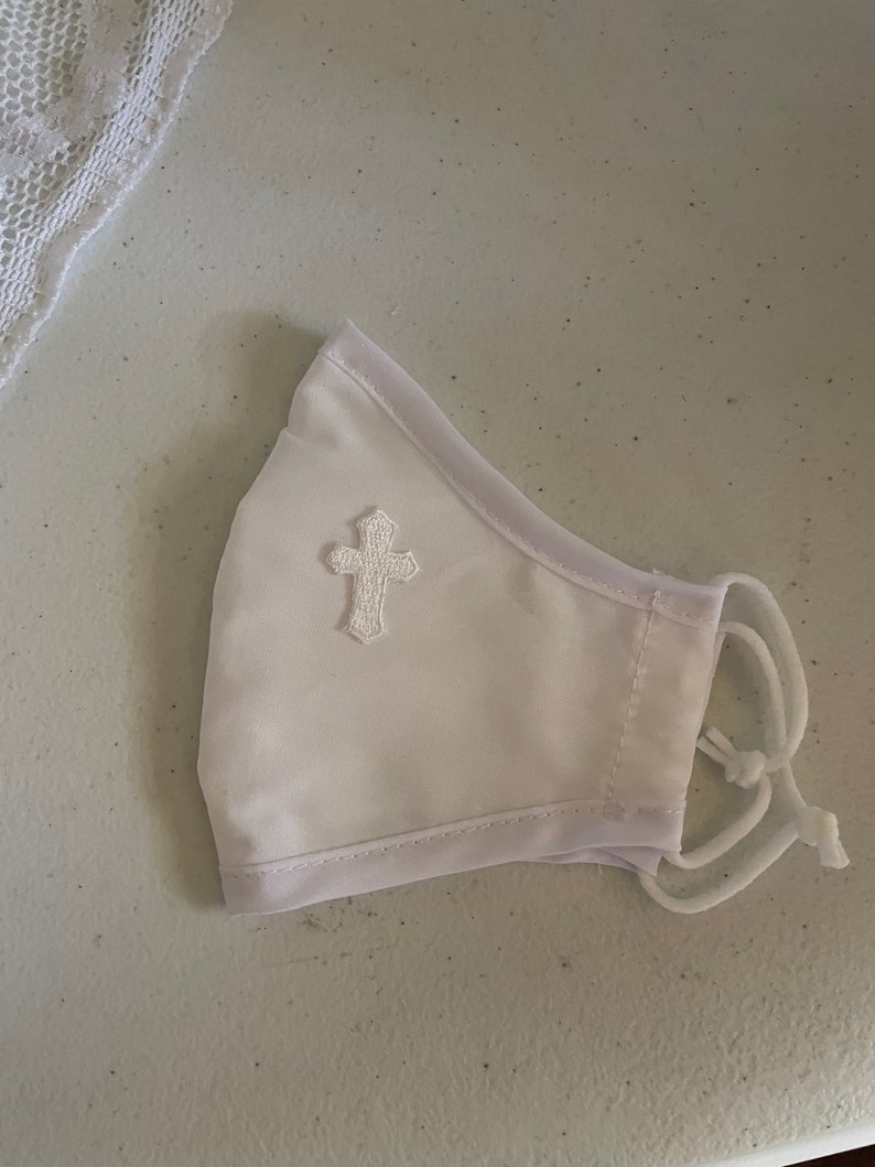 May include: White face mask with a decorative embroidered cross. The mask has adjustable white elastic ear loops and a white trim. A piece of white lace is visible in the upper left corner.