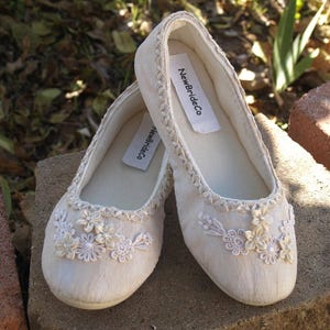 Op de afbeelding: Een paar witte satijnen bruidsslippers met delicate kantversiering en parelversiering. De slippers hebben een label met de tekst "NewBrideCo."