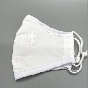 May include: White face mask with a white cross applique on the front. The mask has adjustable white ear loops and is made of a soft, breathable fabric. The mask is designed for comfort and protection.