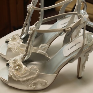 May include: A pair of white satin high-heeled sandals featuring a T-strap design, embellished with intricate white lace, clear rhinestones, and small white pearls. The shoes have an adjustable ankle strap and the brand name NewBrideCo on the insole.
