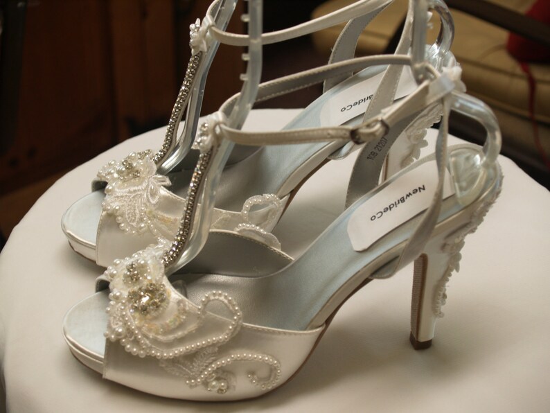 May include: A pair of white satin high-heeled sandals featuring a T-strap design, embellished with intricate white lace, clear rhinestones, and small white pearls. The shoes have an adjustable ankle strap and the brand name NewBrideCo on the insole.