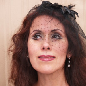 Puede incluir: Una mujer con el pelo largo y castaño lleva un velo negro con una pequeña flor negra y una pluma negra. Lleva un par de pendientes de plata.