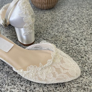 May include: Ivory satin bridal shoes with floral lace overlay and small block heels. The open-toe design features delicate lace detailing and a strap around the ankle. The shoes are embellished with small pearls.