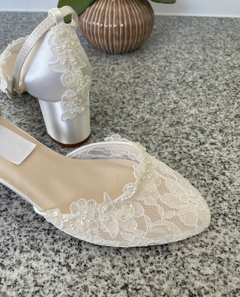 May include: Ivory satin bridal shoes with floral lace overlay and small block heels. The open-toe design features delicate lace detailing and a strap around the ankle. The shoes are embellished with small pearls.