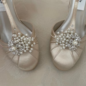 May include: A pair of ivory satin bridal shoes with a t-strap design. Each shoe features a pearl and rhinestone embellishment.