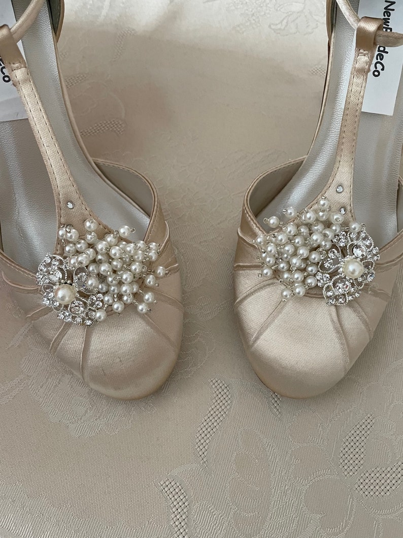 May include: A pair of ivory satin bridal shoes with a t-strap design. Each shoe features a pearl and rhinestone embellishment.