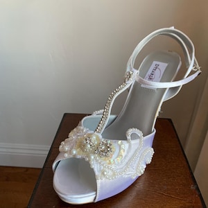 May include: A white satin t-strap high heel shoe with an open toe. The front features intricate white lace, small white pearls, and clear rhinestones. A thin strap with rhinestones runs up the center, and a buckle strap secures the ankle.