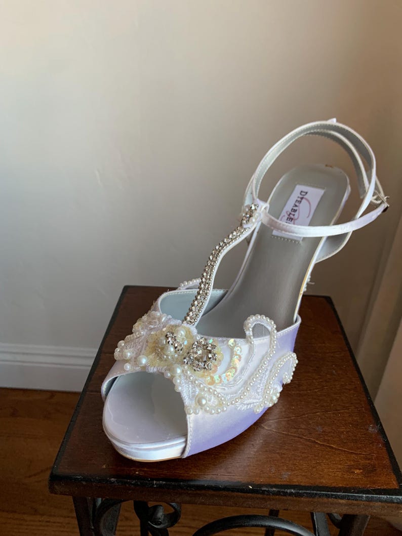 May include: A white satin t-strap high heel shoe with an open toe. The front features intricate white lace, small white pearls, and clear rhinestones. A thin strap with rhinestones runs up the center, and a buckle strap secures the ankle.