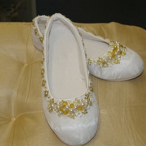 May include: A pair of white satin slippers with gold and pearl embellishments. The slippers have a pointed toe and a low heel.