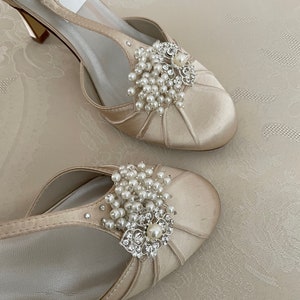May include: A pair of ivory satin bridal shoes with a delicate ankle strap and a pearl and rhinestone embellishment. The shoes have a low heel and a pointed toe.