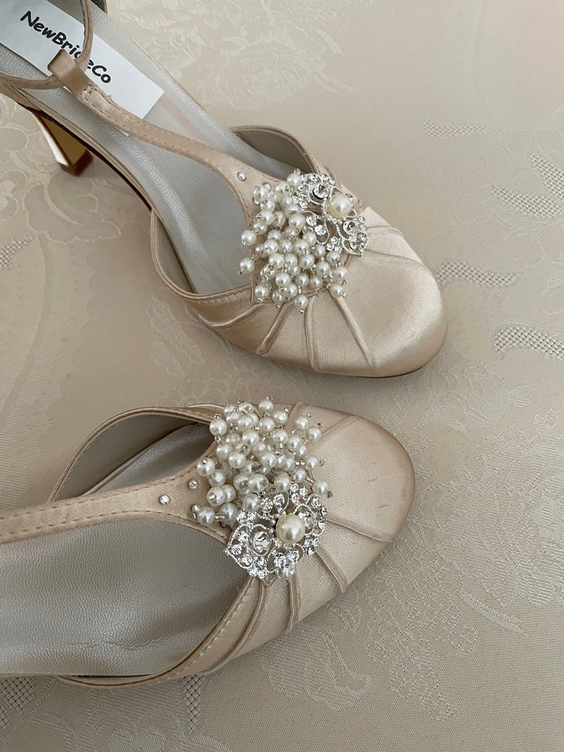 May include: A pair of ivory satin bridal shoes with a delicate ankle strap and a pearl and rhinestone embellishment. The shoes have a low heel and a pointed toe.