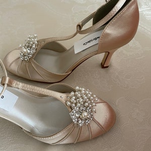 May include: A pair of ivory satin bridal shoes with a t-strap and a pearl and rhinestone embellishment. The shoes have a delicate heel and are labeled "WB Bride Co."
