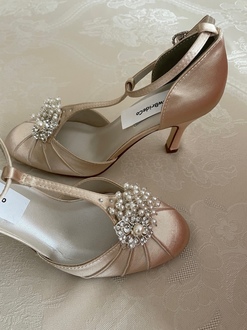 May include: A pair of ivory satin bridal shoes with a t-strap and a pearl and rhinestone embellishment. The shoes have a delicate heel and are labeled "WB Bride Co."