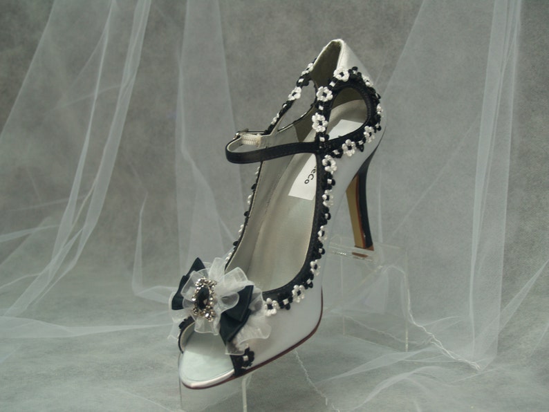 May include: A silver satin high heel shoe with a black bow and floral embellishments. The shoe has a peep toe and a high heel.