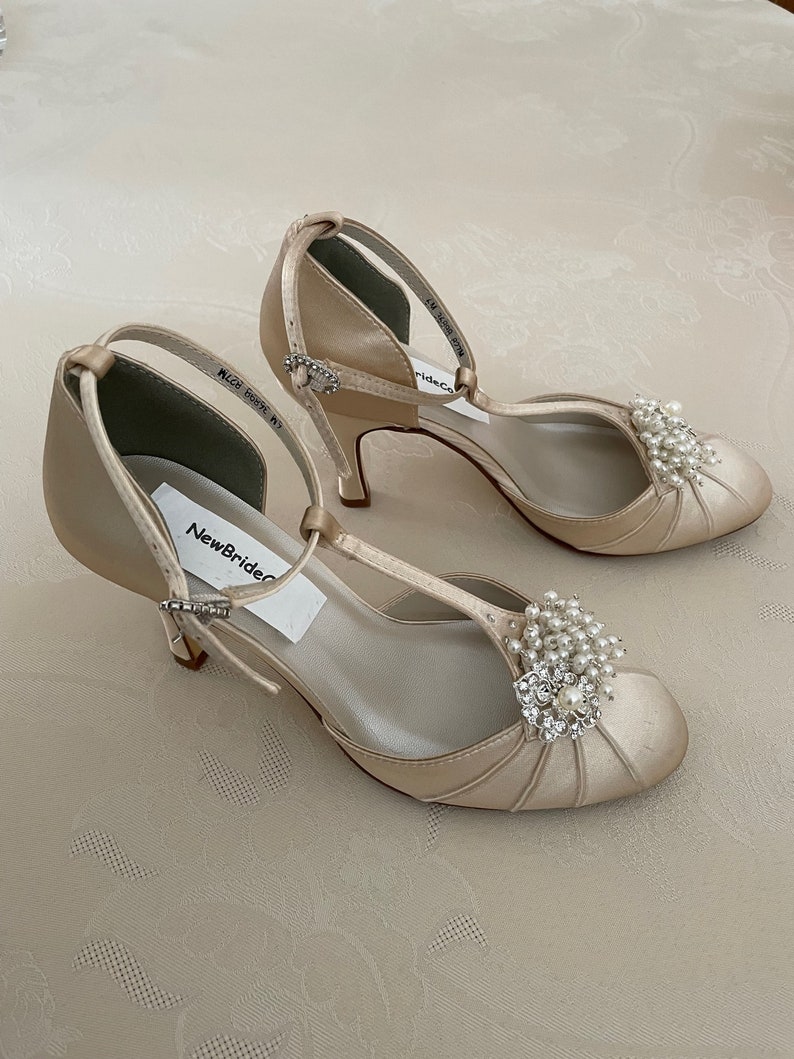 May include: A pair of ivory satin bridal shoes with a t-strap design and a small pearl and rhinestone embellishment. The shoes have a low heel and are labeled "NewBrideCo."