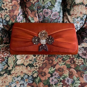May include: A rectangular rust-colored satin clutch bag featuring a central decorative cluster of three fabric flowers, metallic beads, and clear rhinestones. The bag has a smooth, folded fabric texture and a structured, elongated rectangular shape.