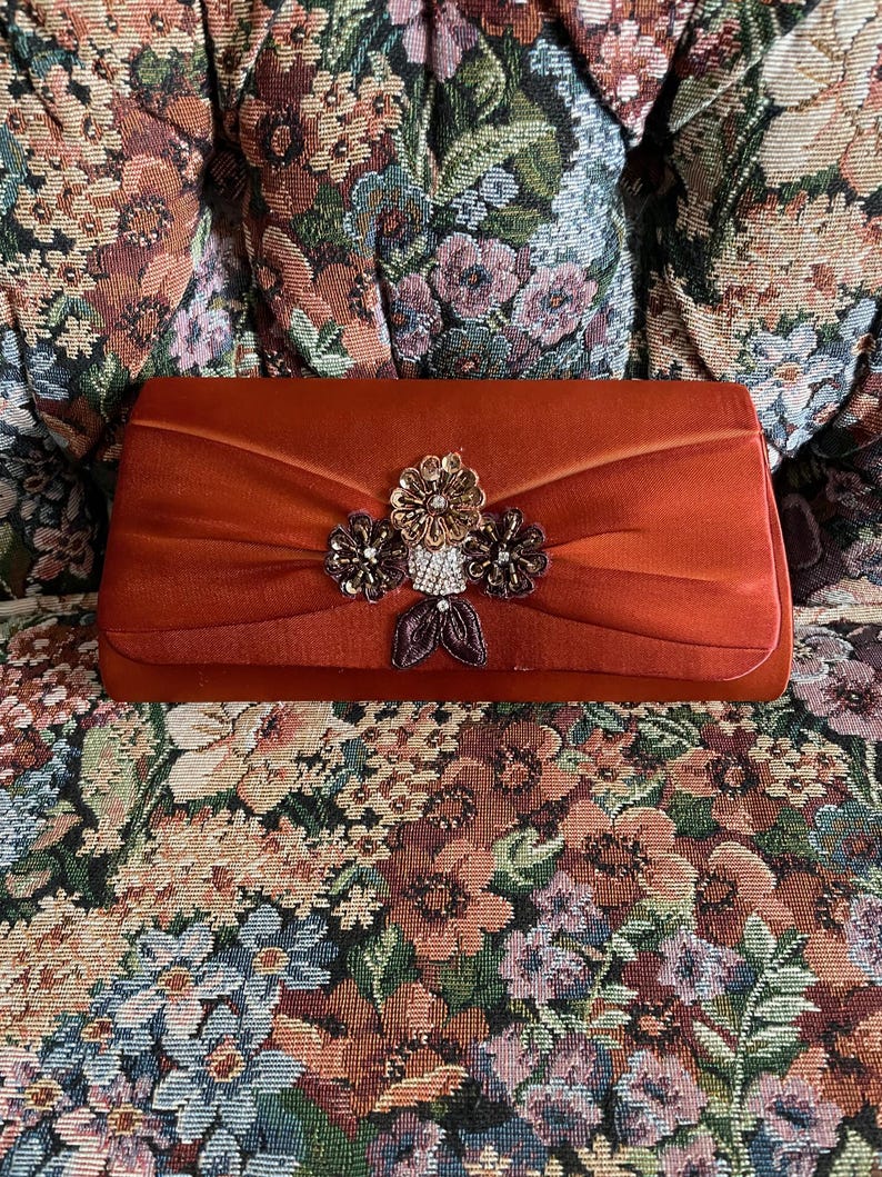 May include: A rectangular rust-colored satin clutch bag featuring a central decorative cluster of three fabric flowers, metallic beads, and clear rhinestones. The bag has a smooth, folded fabric texture and a structured, elongated rectangular shape.
