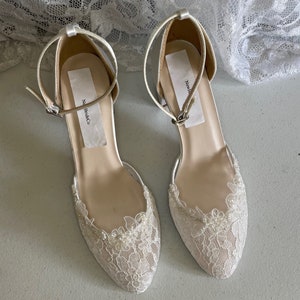 May include: A pair of ivory-colored bridal flats with delicate lace detailing. The shoes feature a closed toe design, ankle straps with buckles, and a low heel. The lace is adorned with small, pearl-like beads. The brand name "New Bride Co" is visible on the insole.