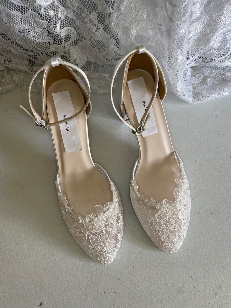 May include: A pair of ivory-colored bridal flats with delicate lace detailing. The shoes feature a closed toe design, ankle straps with buckles, and a low heel. The lace is adorned with small, pearl-like beads. The brand name "New Bride Co" is visible on the insole.