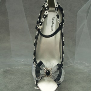 May include: White satin peep-toe heels with black ribbon and beaded embellishments. The heels feature a black bow with a rhinestone centre and the text "NewBrideCo" on the strap.