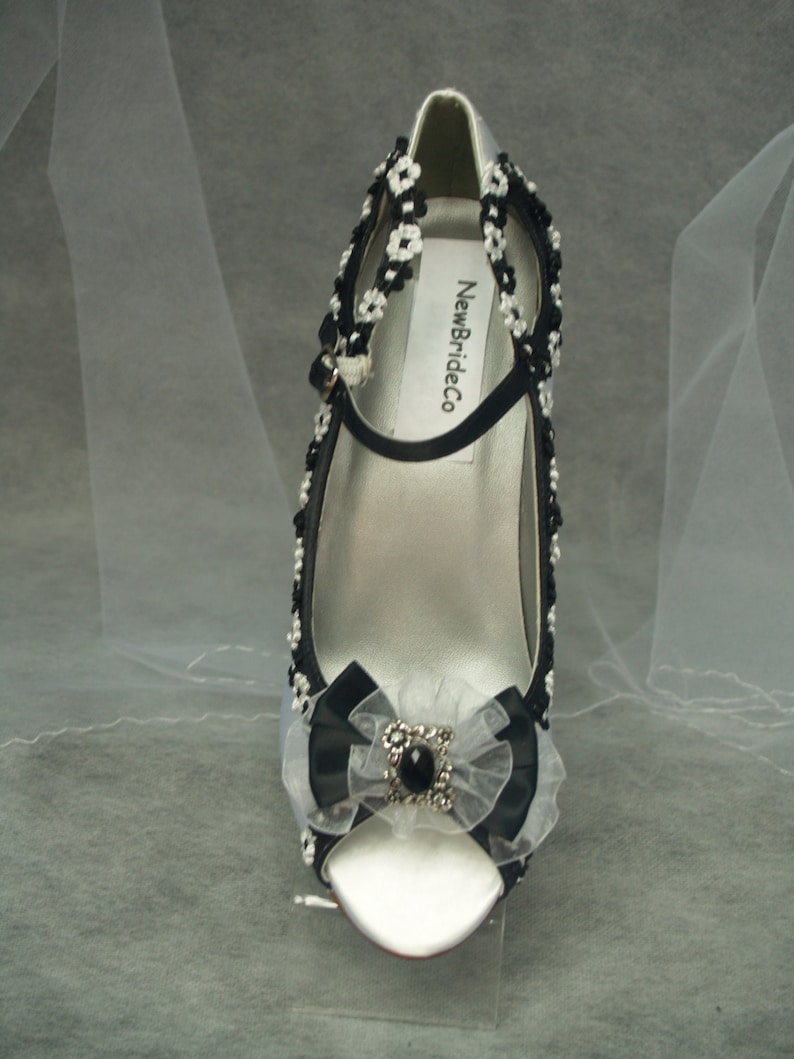 May include: White satin peep-toe heels with black ribbon and beaded embellishments. The heels feature a black bow with a rhinestone centre and the text "NewBrideCo" on the strap.