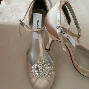 May include: A pair of ivory satin bridal shoes with a T-strap and a pearl and rhinestone embellishment. The shoes have a high heel and a closed toe. The label on the shoe reads "New Bride".