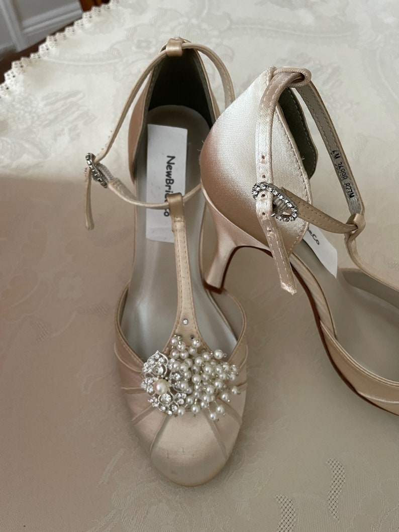 May include: A pair of ivory satin bridal shoes with a T-strap and a pearl and rhinestone embellishment. The shoes have a high heel and a closed toe. The label on the shoe reads "New Bride".