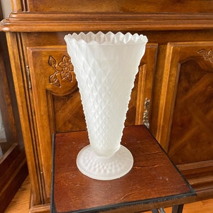 May include: A white frosted glass vase with a diamond pattern and a scalloped rim. The vase is sitting on a wooden table with a black metal base.