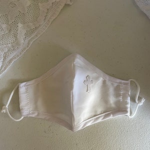 May include: White face mask with a decorative embroidered cross on the front. The mask has adjustable ear loops and is made of a soft, white fabric. A piece of white lace is in the background.