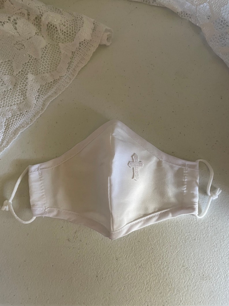 May include: White face mask with a decorative embroidered cross on the front. The mask has adjustable ear loops and is made of a soft, white fabric. A piece of white lace is in the background.