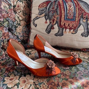 May include: A pair of burnt orange satin kitten heel pumps featuring an open-toe design with a decorative floral appliqu&eacute; made of beads and sequins. The shoes have a side-cut silhouette, a slim heel, and a light-colored interior lining.