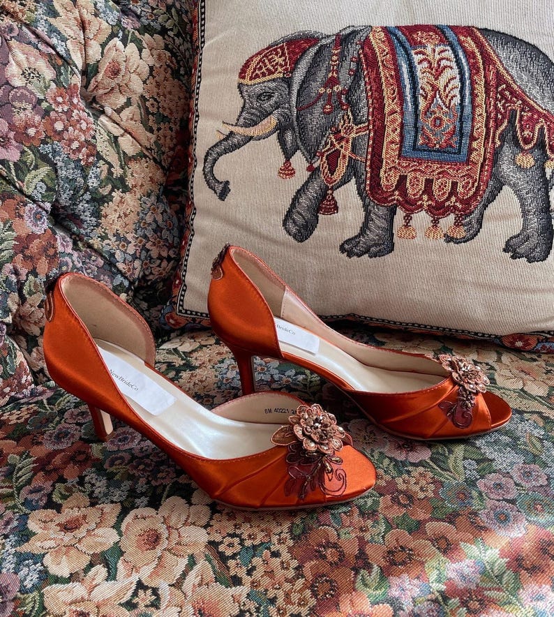 May include: A pair of burnt orange satin kitten heel pumps featuring an open-toe design with a decorative floral appliqu&eacute; made of beads and sequins. The shoes have a side-cut silhouette, a slim heel, and a light-colored interior lining.