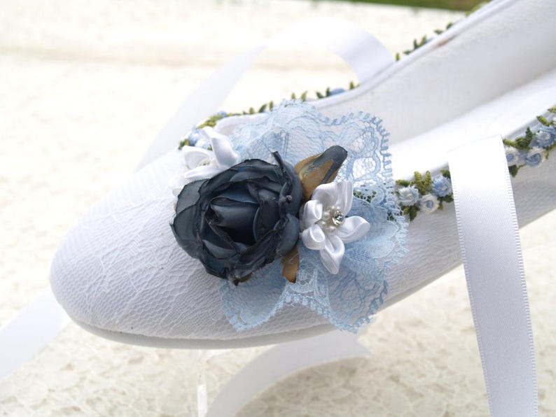 May include: White lace ballet flats with a blue and white flower embellishment. The flower is made of fabric and has a pearl in the center.