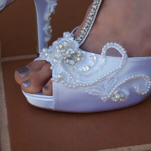 May include: A white satin peep-toe high heel shoe featuring an intricate embellishment of clear crystals, white seed beads, and faux pearls arranged in a swirling pattern across the front, with a crystal-encrusted T-strap detail.