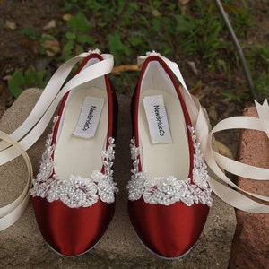 May include: A pair of burgundy satin ballet flats with ivory lace and bead embellishments. The shoes have ivory ribbon ties and the brand name "NewBrideCo" is printed on the insole. The shoes are displayed on a stone surface.