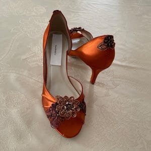 May include: A pair of burnt orange satin open-toe heels featuring a floral appliqu&eacute; with dark brown beads and sequins on the toe. The heel counter is adorned with a matching beaded floral accent. A white label reads New BrideCo inside the shoe.