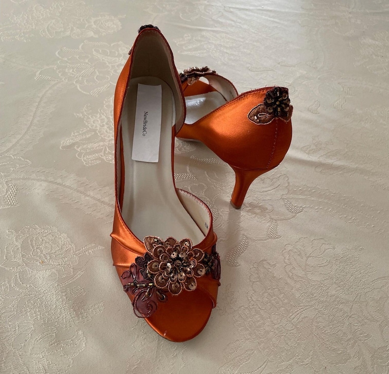 May include: A pair of burnt orange satin open-toe heels featuring a floral appliqu&eacute; with dark brown beads and sequins on the toe. The heel counter is adorned with a matching beaded floral accent. A white label reads New BrideCo inside the shoe.