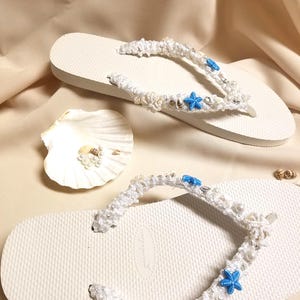 May include: White flip flops with a white crocheted strap decorated with white pearls, blue starfishes, and white seashells.