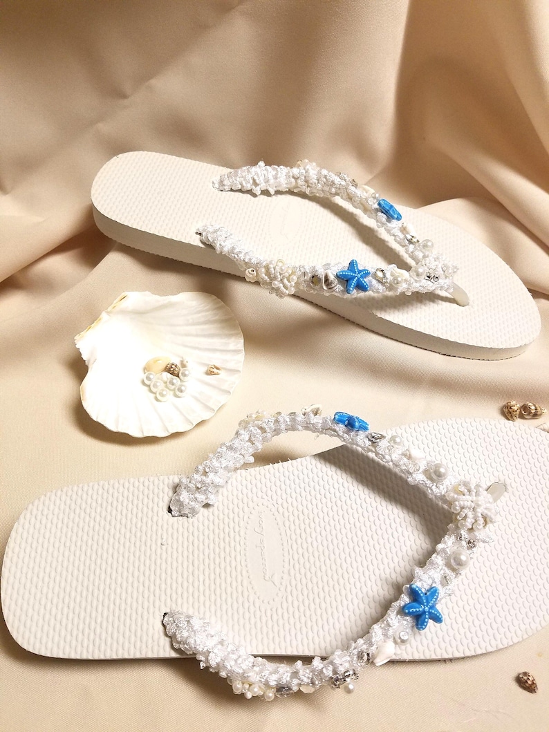 May include: White flip flops with a white crocheted strap decorated with white pearls, blue starfishes, and white seashells.