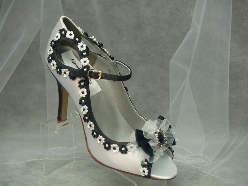May include: White satin bridal shoes with black floral trim and a black bow with a rhinestone embellishment.