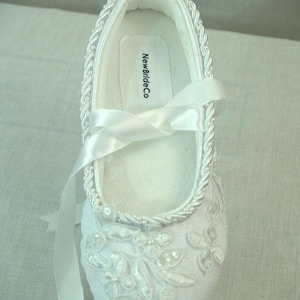 May include: White satin bridal shoes with a delicate floral design and pearl embellishments. The shoes have a ribbon tie and a label that reads "NewBrideCo."