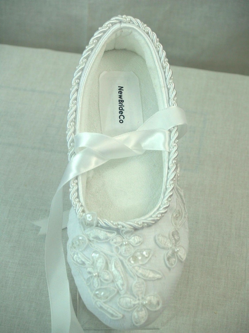 May include: White satin bridal shoes with a delicate floral design and pearl embellishments. The shoes have a ribbon tie and a label that reads "NewBrideCo."