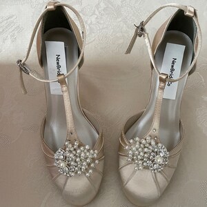 May include: A pair of ivory satin bridal shoes with a T-strap and a pearl and rhinestone embellishment. The shoes have a label that says "NewBridal Co."