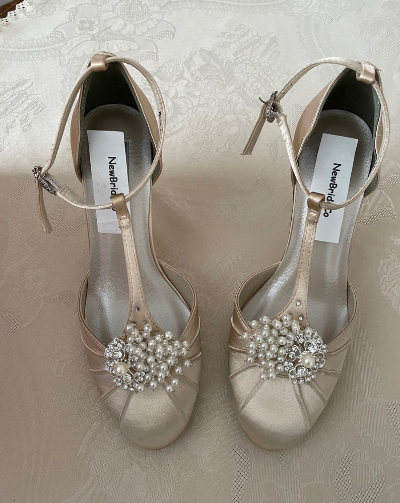 May include: A pair of ivory satin bridal shoes with a T-strap and a pearl and rhinestone embellishment. The shoes have a label that says "NewBridal Co."